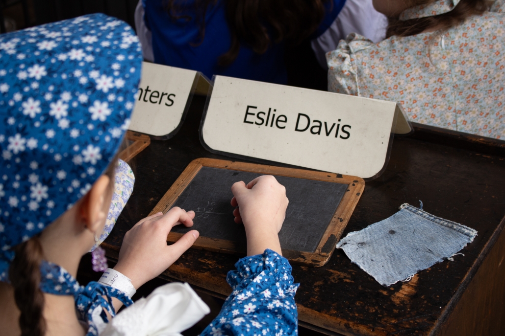 Children dressed in 1800s clothing writes on a slate board on a wooden desk.