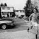 photograph from the 1970s of a young man running in cut-off shorts, smiling in front of a vintage car a d house.