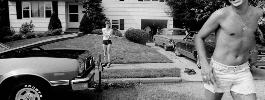 photograph from the 1970s of a young man running in cut-off shorts, smiling in front of a vintage car a d house.