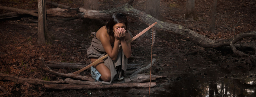Photograph of a young man dressed in Native American regalia drinking from a river in the woods.
